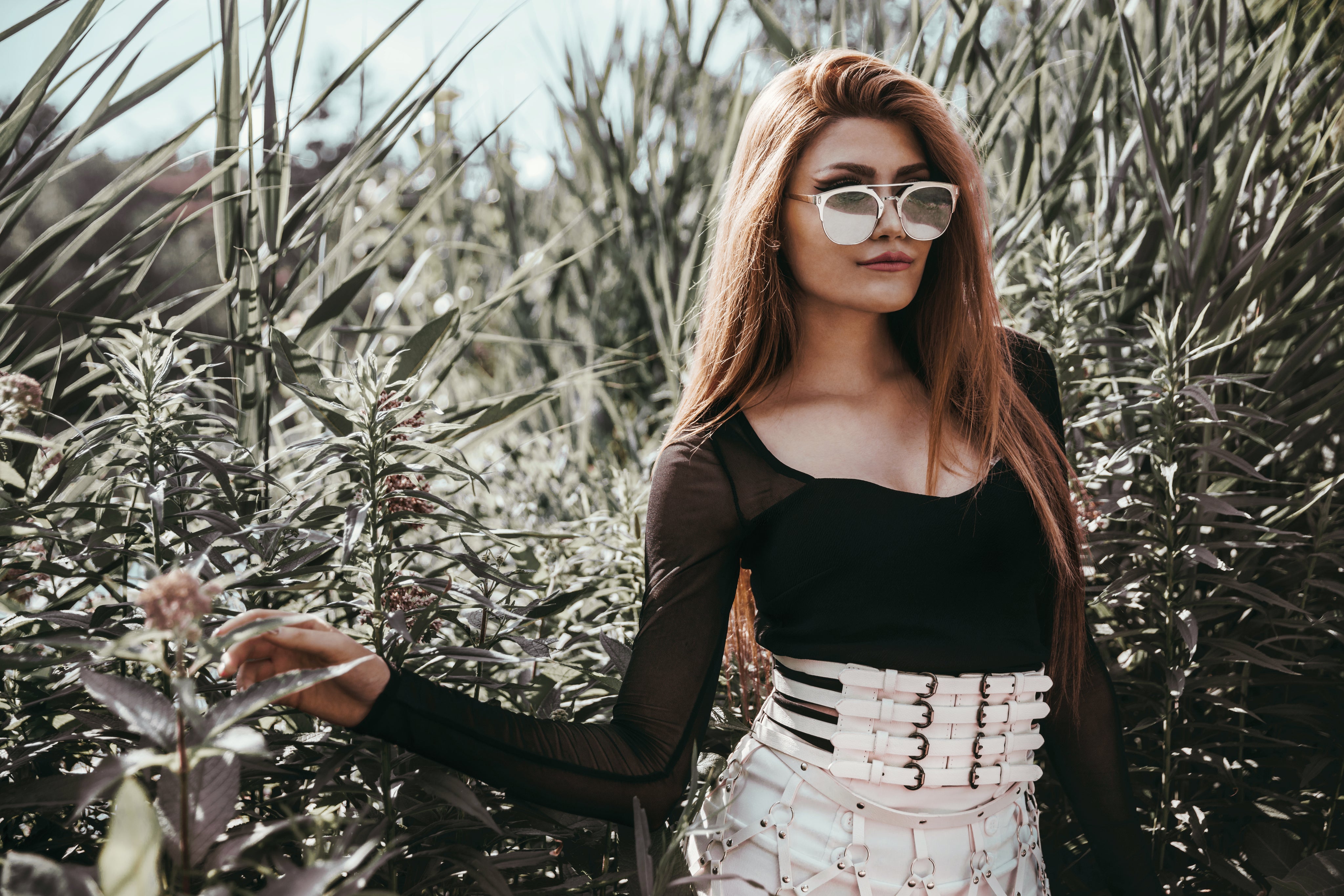 Fashion model in sunglasses and a black top posing outdoors near green bushes